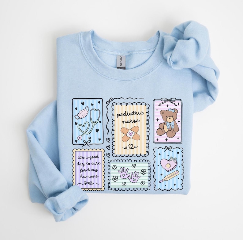 May include: Light blue sweatshirt with a colorful graphic design. The design includes illustrations of a stethoscope, teddy bear, band-aid, and the words "pediatric nurse" and "it's a good day to care for tiny humans."