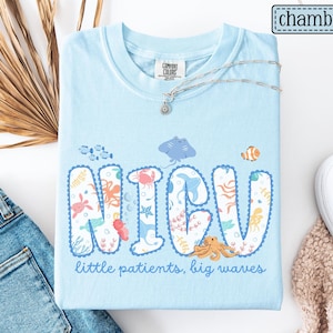 May include: Light blue NICU t-shirt with marine life graphics and the text "NICU little patients, big waves." The shirt features a crew neck and short sleeves. A silver necklace with a pendant is draped across the shirt.