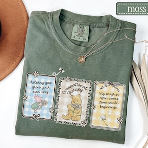 May include: A moss green t-shirt with three framed illustrations featuring Winnie the Pooh characters and inspirational quotes. The shirt has a crew neck and a small Comfort Colors tag. A delicate gold necklace rests on the shirt.