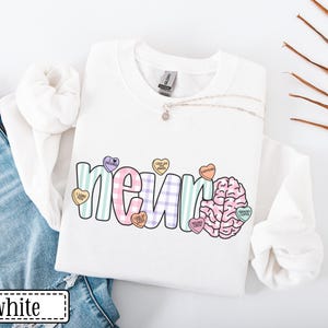 May include: White sweatshirt with the word "neuro" in a colorful, playful font, accompanied by a brain graphic and heart-shaped candies. The candies have phrases like "love on brain" and "caffeine". The word "white" is in a white rectangle.