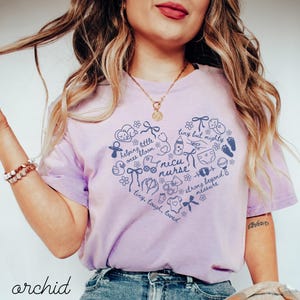 May include: A light purple t-shirt with a heart-shaped design filled with baby-related illustrations and the words "NICU nurse." The shirt also includes the words "helping little ones bloom" and "tiny but mighty."