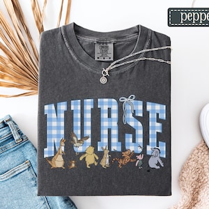 K&ouml;nnte beinhalten: Dunkelgraues T-Shirt mit dem Wort "NURSE" in blau-wei&szlig; karierten Buchstaben. Darunter befinden sich Zeichentrickfiguren. Eine silberne Halskette liegt &uuml;ber dem Shirt. Der Markenname "pepper" steht in einem gr&uuml;nen Rechteck.