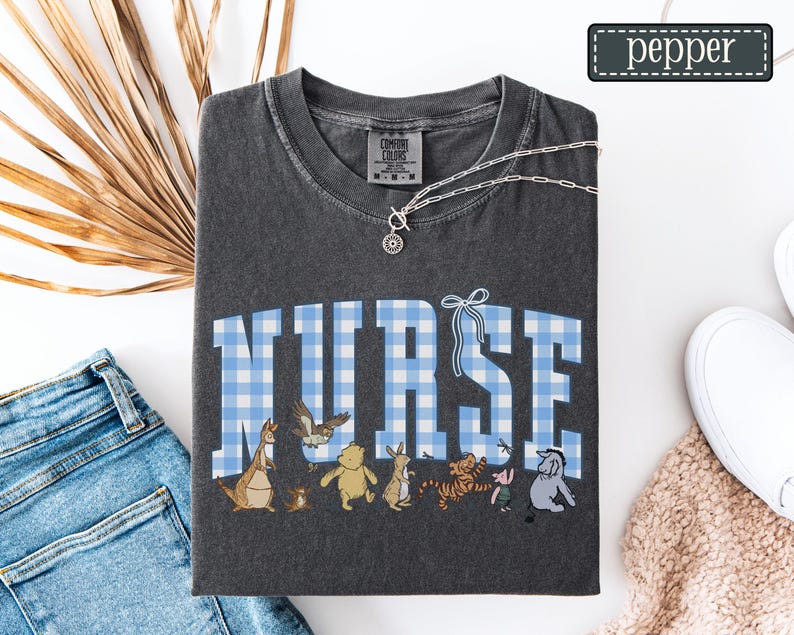 K&ouml;nnte beinhalten: Dunkelgraues T-Shirt mit dem Wort "NURSE" in blau-wei&szlig; karierten Buchstaben. Darunter befinden sich Zeichentrickfiguren. Eine silberne Halskette liegt &uuml;ber dem Shirt. Der Markenname "pepper" steht in einem gr&uuml;nen Rechteck.