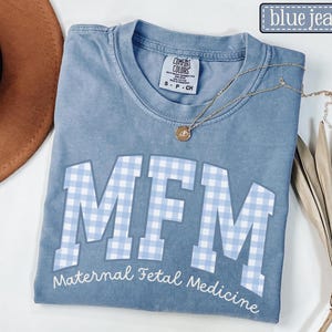 May include: A blue jean colored t-shirt with the letters "MFM" in white, filled with a light blue gingham pattern. The words "Maternal Fetal Medicine" are printed below the letters. A gold necklace is draped across the shirt.