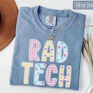 May include: A blue jean Comfort Colors t-shirt with the words "RAD TECH" in a colorful, patterned design. The letters are decorated with gingham and floral patterns. A gold necklace with a pendant is draped across the shirt.