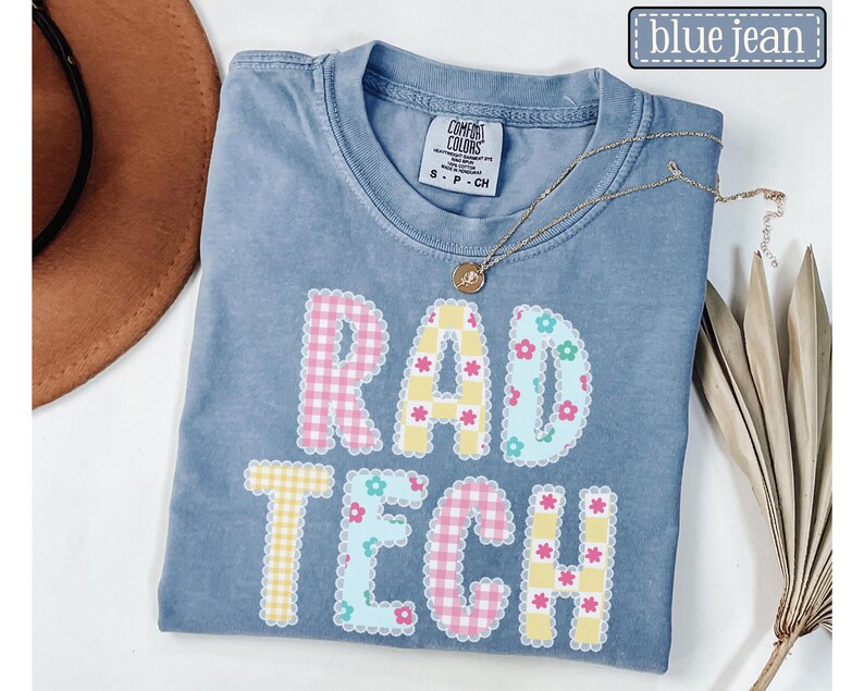 May include: A blue jean Comfort Colors t-shirt with the words "RAD TECH" in a colorful, patterned design. The letters are decorated with gingham and floral patterns. A gold necklace with a pendant is draped across the shirt.