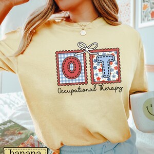 May include: A light yellow t-shirt featuring the letters "OT" in a postage stamp design. The "O" is red with a white and blue gingham pattern, and the "T" is blue with red and white floral accents. The words "Occupational Therapy" are printed below.