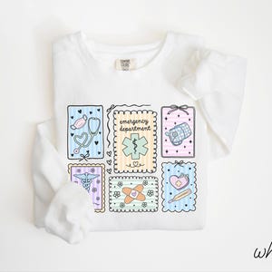 Pode incluir: Camisola branca de manga comprida com um design de tema m&eacute;dico. O design inclui um estetosc&oacute;pio, uma mala m&eacute;dica e outros s&iacute;mbolos m&eacute;dicos. As palavras "emergency department" tamb&eacute;m est&atilde;o inclu&iacute;das.