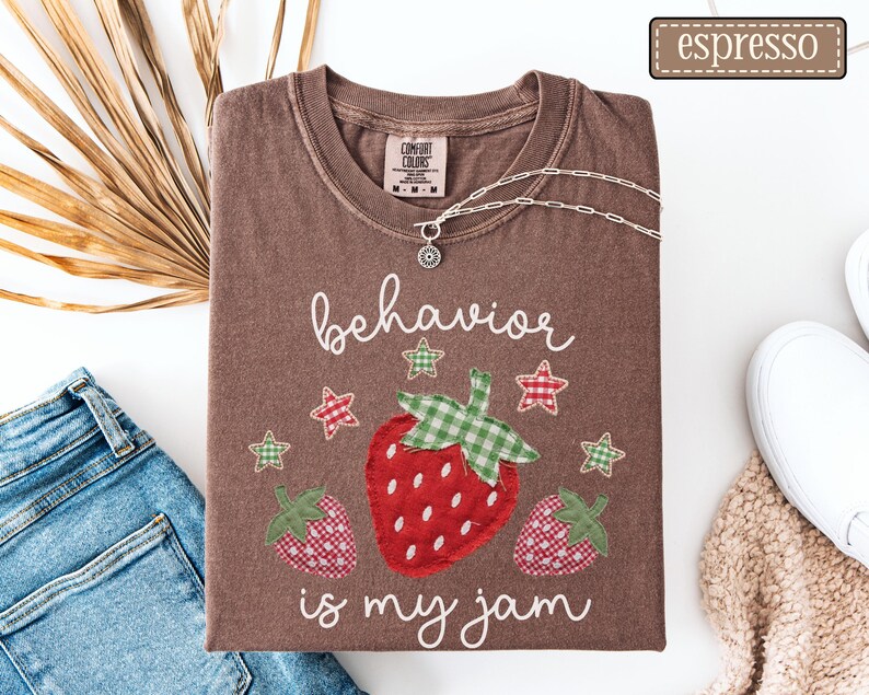 K&ouml;nnte beinhalten: Ein espressofarbenes T-Shirt mit einer Grafik von drei Erdbeeren in rot-gr&uuml;nen Vichy-Karomustern. Der Text "behavior is my jam" ist in wei&szlig;er Schreibschrift gedruckt, erg&auml;nzt durch kleine rot-gr&uuml;ne Vichy-Sterne.