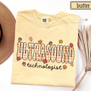 Puede incluir: Una camiseta amarilla clara con la palabra "ULTRASOUND" en un dise&ntilde;o a cuadros y la palabra "technologist" debajo. La camiseta tiene gr&aacute;ficos de hojas y bellotas con tem&aacute;tica oto&ntilde;al. La camiseta est&aacute; etiquetada como "butter".