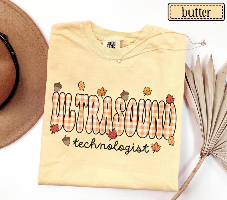 Puede incluir: Una camiseta amarilla clara con la palabra "ULTRASOUND" en un dise&ntilde;o a cuadros y la palabra "technologist" debajo. La camiseta tiene gr&aacute;ficos de hojas y bellotas con tem&aacute;tica oto&ntilde;al. La camiseta est&aacute; etiquetada como "butter".