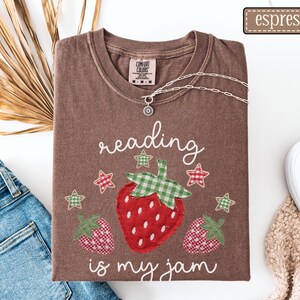May include: An espresso brown short-sleeve t-shirt featuring a graphic of three strawberries with green gingham leaves and red gingham patterns, surrounded by small stars and the white text reading, reading is my jam.