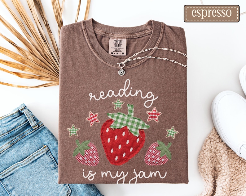 May include: An espresso brown short-sleeve t-shirt featuring a graphic of three strawberries with green gingham leaves and red gingham patterns, surrounded by small stars and the white text reading, reading is my jam.