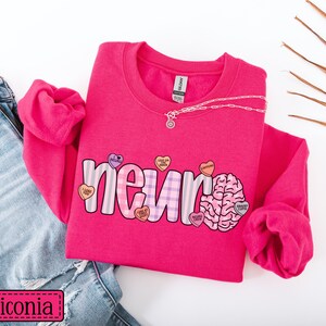May include: A bright pink sweatshirt with the word "neuro" in a playful font, accented with a brain graphic and heart-shaped candies. The sweatshirt is paired with blue jeans and a delicate necklace.