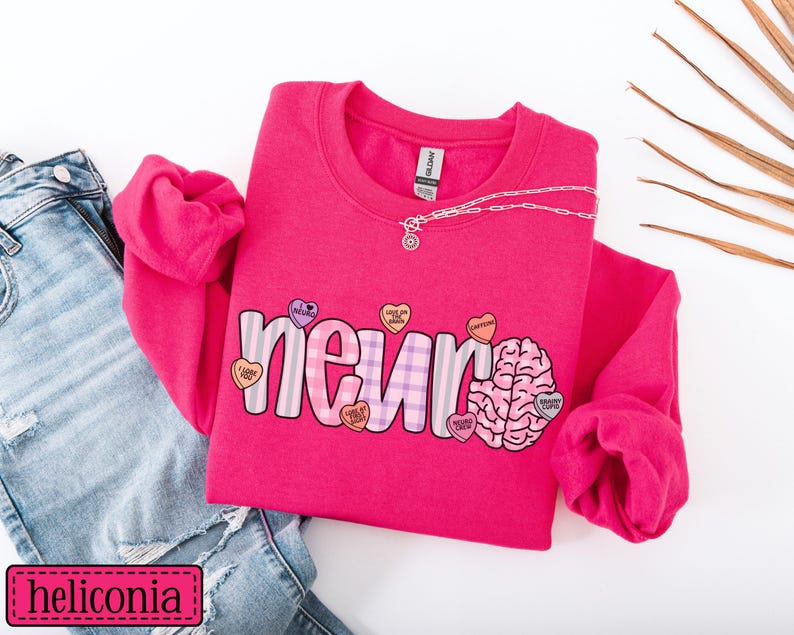 May include: A bright pink sweatshirt with the word "neuro" in a playful font, accented with a brain graphic and heart-shaped candies. The sweatshirt is paired with blue jeans and a delicate necklace.