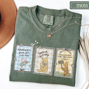 May include: A moss green t-shirt with three framed illustrations featuring Winnie the Pooh characters. The illustrations include text that reads "helping you grow your own way," "Speech Therapy," and "Communication looks different for everyone."