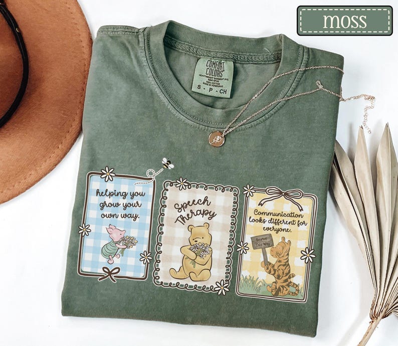 May include: A moss green t-shirt with three framed illustrations featuring Winnie the Pooh characters. The illustrations include text that reads "helping you grow your own way," "Speech Therapy," and "Communication looks different for everyone."