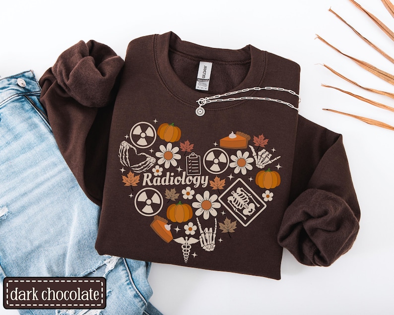 May include: A dark chocolate brown crewneck sweatshirt featuring a heart-shaped graphic composed of pumpkins, daisies, maple leaves, radiation symbols, a clipboard, a skeleton hand, a slice of pie, and a skeleton rib cage with the word Radiology.
