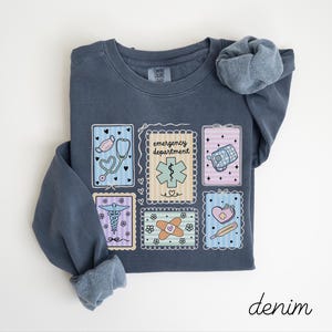 Pode incluir: Uma camisa de manga comprida azul denim com gola redonda. A camisa apresenta um design de ilustra&ccedil;&otilde;es com tema m&eacute;dico dentro de molduras decorativas, incluindo um estetosc&oacute;pio, uma m&aacute;scara e um term&oacute;metro. A palavra "emergency department" tamb&eacute;m &eacute; exibida.