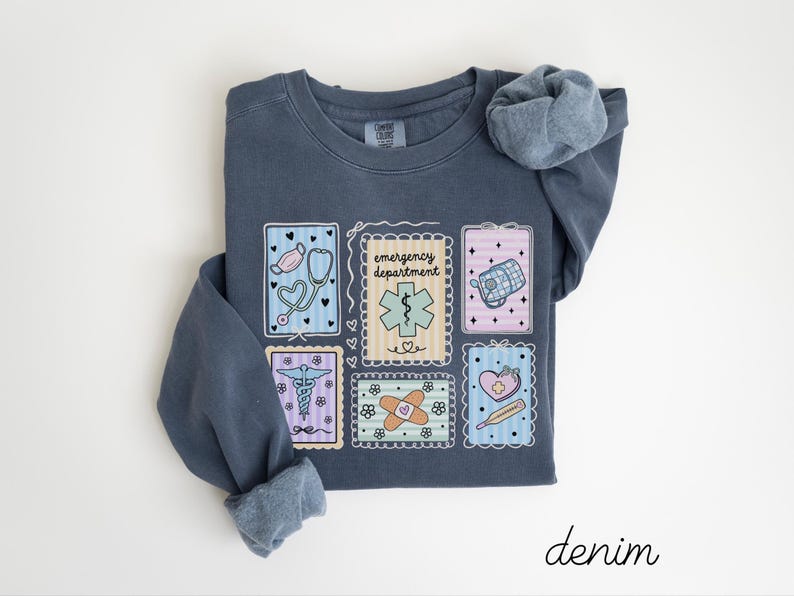 Pode incluir: Uma camisa de manga comprida azul denim com gola redonda. A camisa apresenta um design de ilustra&ccedil;&otilde;es com tema m&eacute;dico dentro de molduras decorativas, incluindo um estetosc&oacute;pio, uma m&aacute;scara e um term&oacute;metro. A palavra "emergency department" tamb&eacute;m &eacute; exibida.