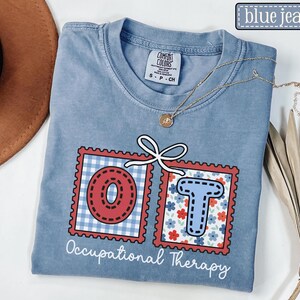 May include: A blue jean Comfort Colors t-shirt with the words "OT" in red and blue postage stamp designs. The "O" is red with a white gingham background, and the "T" is blue with a floral background. The words "Occupational Therapy" are printed below.