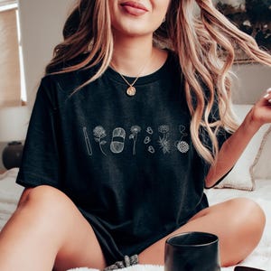 May include: A black t-shirt with a white graphic design of a crochet hook, yarn, and flowers. The graphic design is centered on the shirt.