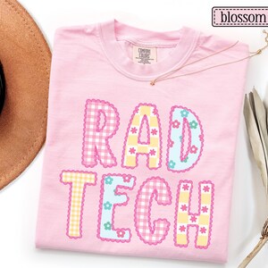 May include: A light pink t-shirt with the words "RAD TECH" in a colorful, playful design. The letters are decorated with gingham patterns, flowers, and scalloped edges. The word "blossom" is in a small box in the upper right corner.