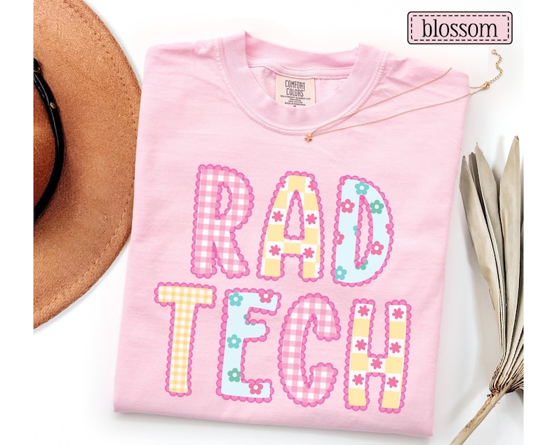 May include: A light pink t-shirt with the words "RAD TECH" in a colorful, playful design. The letters are decorated with gingham patterns, flowers, and scalloped edges. The word "blossom" is in a small box in the upper right corner.