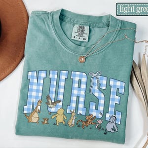 K&ouml;nnte beinhalten: Ein hellgr&uuml;nes T-Shirt mit dem Wort "NURSE" in blau-karierten Buchstaben. Das Shirt zeigt Zeichentrickfiguren unter dem Wort. Eine goldene Halskette mit Anh&auml;nger befindet sich auf dem Shirt.