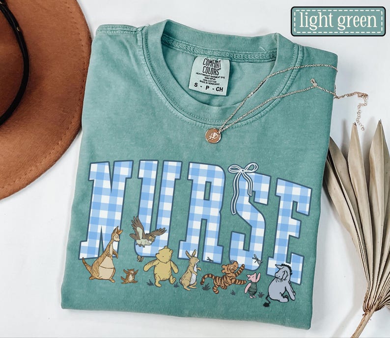 K&ouml;nnte beinhalten: Ein hellgr&uuml;nes T-Shirt mit dem Wort "NURSE" in blau-karierten Buchstaben. Das Shirt zeigt Zeichentrickfiguren unter dem Wort. Eine goldene Halskette mit Anh&auml;nger befindet sich auf dem Shirt.