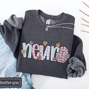 May include: Dark heather gray sweatshirt with the word "neuro" in pastel colors and a brain graphic. The sweatshirt has a silver necklace and is paired with blue jeans. The word "dark heather gray" is in a black rectangle.