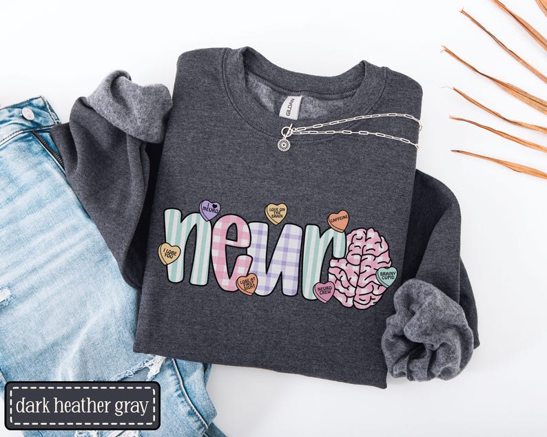 May include: Dark heather gray sweatshirt with the word "neuro" in pastel colors and a brain graphic. The sweatshirt has a silver necklace and is paired with blue jeans. The word "dark heather gray" is in a black rectangle.