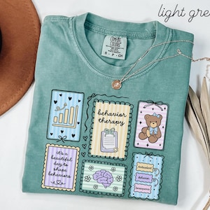 May include: Light green t-shirt with multiple graphic designs. The designs include the words "behavior therapy", "it's a beautiful day to shape behaviors", and a teddy bear. A silver necklace is draped across the shirt.