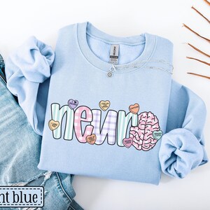 May include: Light blue sweatshirt with the word "neuron" in a colorful, playful font, accompanied by a brain graphic and candy heart accents. The text "light blue" is in a white box at the bottom left.