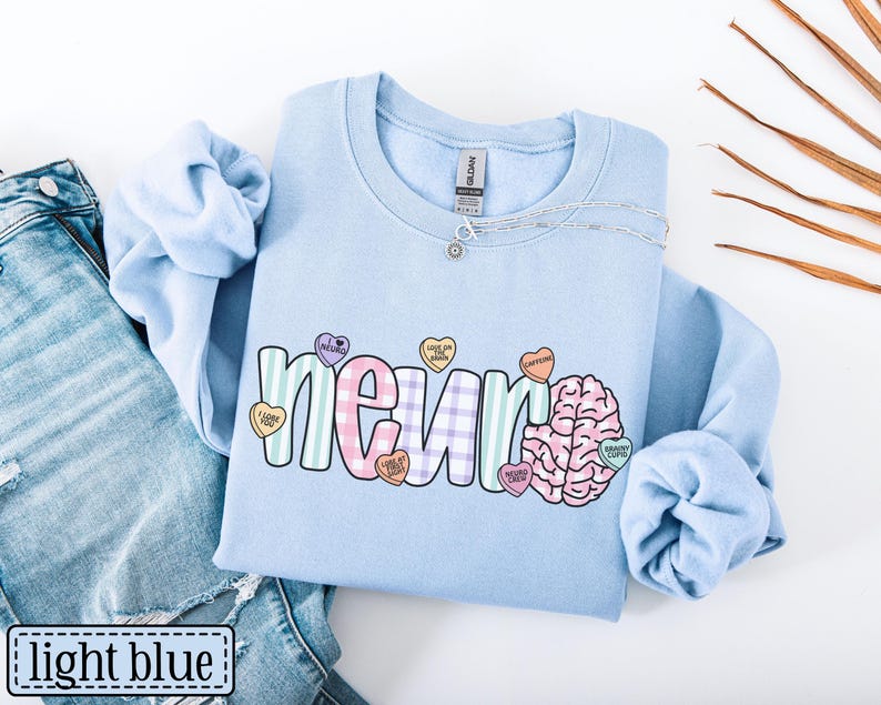 May include: Light blue sweatshirt with the word "neuron" in a colorful, playful font, accompanied by a brain graphic and candy heart accents. The text "light blue" is in a white box at the bottom left.