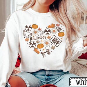 May include: A white long-sleeve sweatshirt featuring a heart-shaped graphic composed of various icons including pumpkins, autumn leaves, daisies, radiation symbols, a clipboard, a slice of pie, a skeleton hand, and the word Radiology in brown.