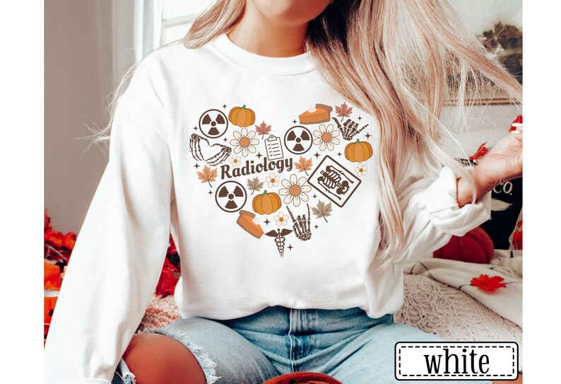 May include: A white long-sleeve sweatshirt featuring a heart-shaped graphic composed of various icons including pumpkins, autumn leaves, daisies, radiation symbols, a clipboard, a slice of pie, a skeleton hand, and the word Radiology in brown.