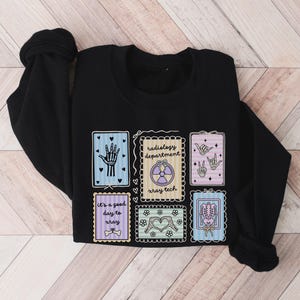 May include: Black sweatshirt featuring colorful illustrations related to radiology. Designs include a hand, a radiation symbol, hand gestures, a skeleton, and the text "It's a good day to xray" and "radiology department xray tech".