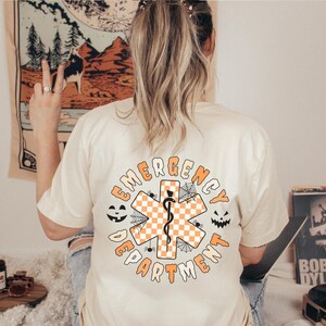 Op de afbeelding: Een cr&egrave;mekleurig t-shirt met een afbeelding op de achterkant met de tekst EMERGENCY DEPARTMENT in het oranje. Het ontwerp bevat een centraal oranje en wit geblokt symbool van de Ster van het Leven met een zwarte slang en spinnenwebben.