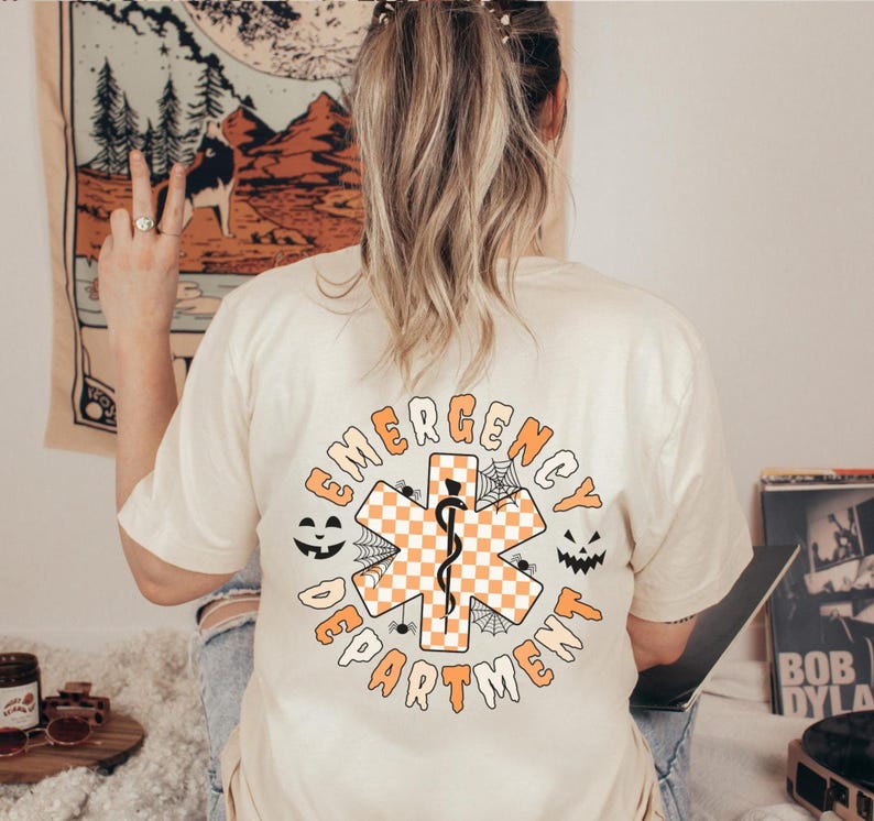 Op de afbeelding: Een cr&egrave;mekleurig t-shirt met een afbeelding op de achterkant met de tekst EMERGENCY DEPARTMENT in het oranje. Het ontwerp bevat een centraal oranje en wit geblokt symbool van de Ster van het Leven met een zwarte slang en spinnenwebben.