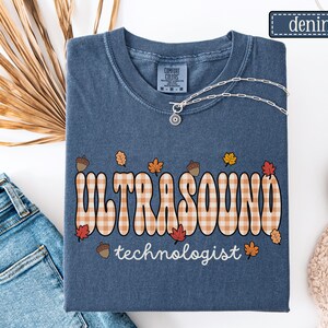Puede incluir: Camiseta azul denim con las palabras "ULTRASOUND technologist" en una fuente retro con un patr&oacute;n de cuadros. El dise&ntilde;o incluye hojas de oto&ntilde;o y bellotas. Un collar plateado con un colgante est&aacute; sobre la camiseta.