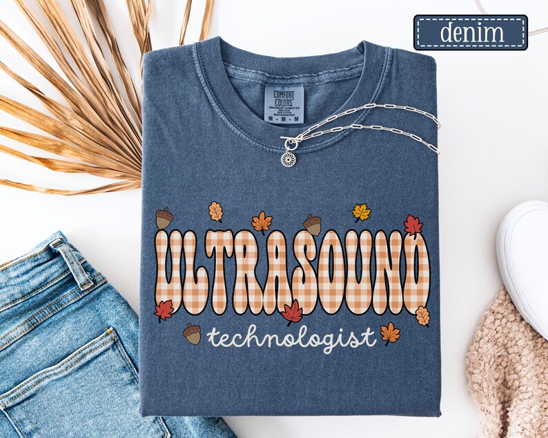 Puede incluir: Camiseta azul denim con las palabras "ULTRASOUND technologist" en una fuente retro con un patr&oacute;n de cuadros. El dise&ntilde;o incluye hojas de oto&ntilde;o y bellotas. Un collar plateado con un colgante est&aacute; sobre la camiseta.
