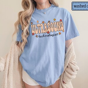 Puede incluir: Camiseta azul claro con las palabras "ULTRASOUND technologist" en un dise&ntilde;o a cuadros con hojas de oto&ntilde;o y bellotas. La camiseta est&aacute; etiquetada como "washed denim".