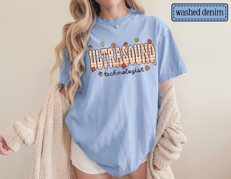 Puede incluir: Camiseta azul claro con las palabras "ULTRASOUND technologist" en un dise&ntilde;o a cuadros con hojas de oto&ntilde;o y bellotas. La camiseta est&aacute; etiquetada como "washed denim".