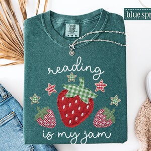 May include: A dark green t-shirt featuring a graphic of three strawberries with green and red checkered patterns. The text reading is my jam is printed in white script above and below the strawberries, accompanied by small checkered stars.