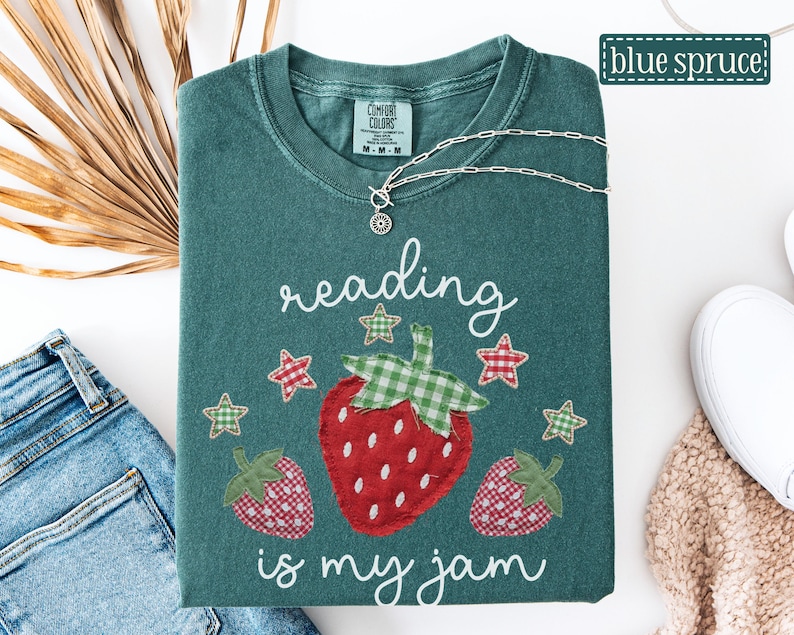 May include: A dark green t-shirt featuring a graphic of three strawberries with green and red checkered patterns. The text reading is my jam is printed in white script above and below the strawberries, accompanied by small checkered stars.