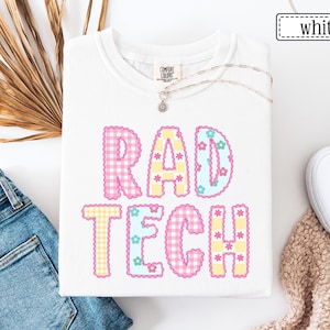 May include: White t-shirt with the text "RAD TECH" in colorful, patterned letters. The letters are pink, yellow, and blue with floral accents. The shirt is folded, and a necklace with a small pendant is visible.