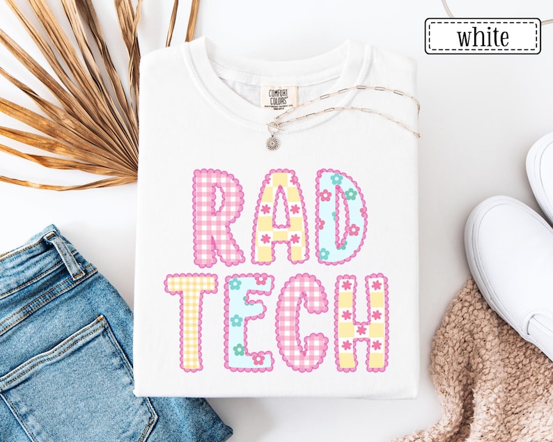 May include: White t-shirt with the text "RAD TECH" in colorful, patterned letters. The letters are pink, yellow, and blue with floral accents. The shirt is folded, and a necklace with a small pendant is visible.