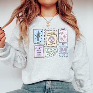 May include: Light gray sweatshirt featuring colorful medical-themed illustrations. Designs include a hand, a heart, a skeleton, and a radiology symbol. Text on the sweatshirt reads "Radiology Department" and "It's a good day to xray."