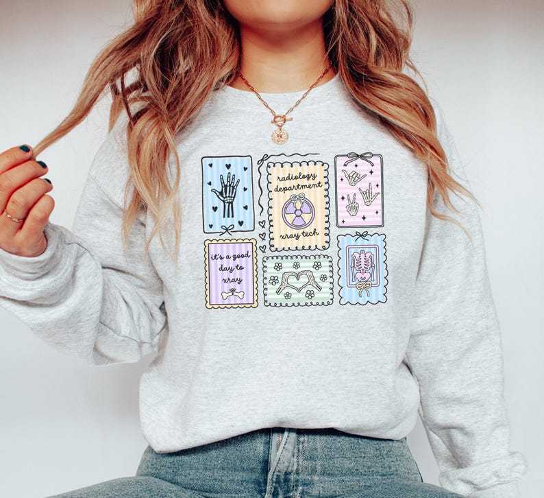 May include: Light gray sweatshirt featuring colorful medical-themed illustrations. Designs include a hand, a heart, a skeleton, and a radiology symbol. Text on the sweatshirt reads "Radiology Department" and "It's a good day to xray."